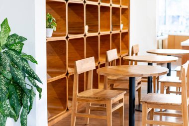 Ahşap masalar ve sandalyelerin bulunduğu rahat bir kafe arka planda şık bir ahşap raf. Yeşil bir bitki uzaya doğanın bir dokunuşunu ekler ve sıcak bir ortam yaratır.