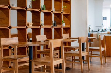 İçinde ahşap masalar ve sandalyeler olan modern bir kafe. Arka planda dekoratif eşyalar ve bitkilerle süslü şık bir ahşap raf ünitesi sergileniyor. Burası çok aydınlık.