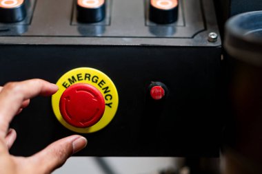 Kontrol panelindeki büyük kırmızı acil durdurma düğmesine uzanan bir elin yakın çekimi. Düğme sarı arkaplanlı daireseldir ve 