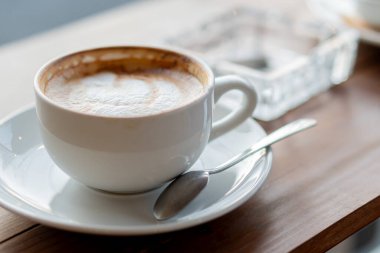 Ahşap bir masaya konmuş, latte sanatıyla dolu beyaz bir kahve fincanının yakın çekimi. Fincana küçük bir kaşık ve arka planda cam bir kül tablası eşlik eder.