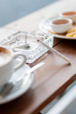 İçinde bir fincan kahve, bir kül tablası, bir sigara ve bir tabak atıştırmalık bulunan rahat bir kafe sahnesi. Ortam sıcak ve davetkar, rahatlamak ya da sohbet etmek için mükemmel..