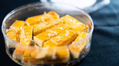 Üzerinde hindistancevizi parçalanmış sarı bir tatlının dikdörtgen şeklinde parçalarıyla dolu şeffaf bir kabın yakın çekim görüntüsü. Tatlı tatlı ve nemli görünüyor, ince zeka.