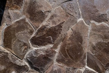 Düzensiz şekilli kahverengi taşlardan oluşan ve gözle görülür çatlaklar ve renk çeşitliliğine sahip dokulu taş bir duvarın yakından görüntüsü. Yüzey doğal ve engebeli görünüyor.