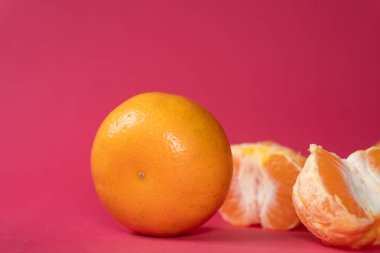 Canlı pembe arka planda iki portakal, bir portakal kısmen soyulmuş, sulu içini gösteriyor. Parlak renkler çarpıcı bir zıtlık yaratır, tazelik ve dokuya vurgu yapar.