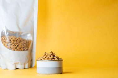 A bowl filled with dry pet food next to a resealable bag on a yellow background. clipart