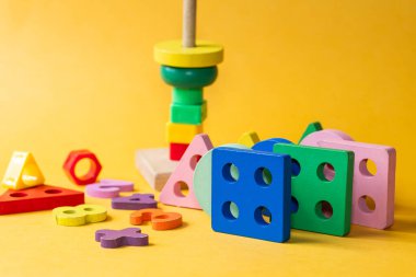 Parlak sarı arka planda, ahşap geometrik oyuncaklardan oluşan renkli bir düzenleme. Oyuncaklar kareler, üçgenler ve renkli daireler gibi çeşitli şekilleri içerir.