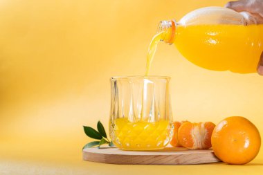 Şişeden portakal suyu dolduran bir elin temiz bir bardağa doldurduğu ferahlatıcı bir sahne. Arka plan canlı bir sarı, meyve suyunun parlak rengini arttırıyor. Dilimlenmiş bir portakal