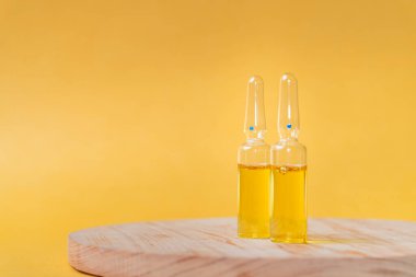 Sarı sıvı içeren iki cam ampul, yumuşak sarı arka planlı ahşap bir yüzeye yerleştirilmiş. Ampullerin üzerinde mavi bir nokta var. Bu da cilt bakımı için bir serum ya da yağ içerdiğini gösteriyor.
