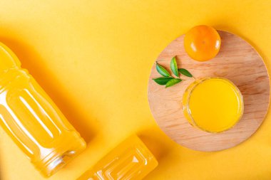 Bir bardak portakal suyu, bir şişe portakal suyu ve ahşap bir tabakta yeşil yapraklı taze bir portakal içeren canlı sarı bir arka plan..