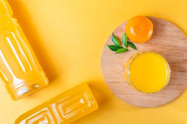 Bir bardak portakal suyu, bir şişe portakal suyu ve ahşap bir tabakta yeşil yapraklı taze bir portakal içeren canlı sarı bir arka plan..