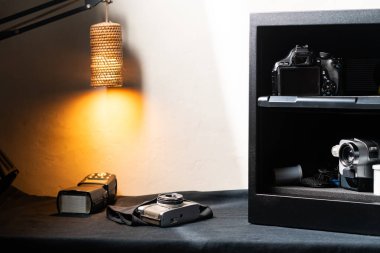 A black electronic safe with a transparent door, containing a camera and a lens inside. The safe is placed on a dark surface with a warm light illuminating the scene from the left. A key is inserted clipart