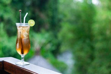 Limon dilimi ve iki pipetle süslenmiş uzun bir bardakta servis edilen ferahlatıcı bir kokteyl. İçki, yemyeşil arka planı olan ahşap bir korkuluğa yerleştirilmiş.