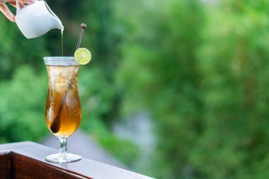 Küçük bir sürahide buzla dolu uzun bir bardağa serinletici bir kokteyl dökülüyor. İçkinin koyu kehribar rengi var ve limon dilimi ve karıştırıcı ile süslenmiş.