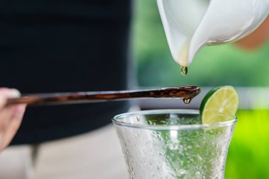 A close-up of a drink preparation scene, featuring a wooden stirrer dripping liquid into a glass. A slice of lime is placed on the rim of the glass clipart