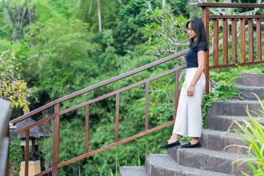 Yeşilliklerle çevrili taş bir merdivende yürüyen bir kadın. Merdiven ahşap bir korkulukla çevrili ve yanında geleneksel bir fener var. Sahne huzur veriyor.