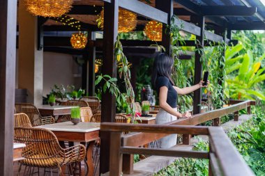 Tropik bir restoran ortamında kendini alan bir kadın. Açık havada ahşap mobilyalar, bereketli yeşillik ve dekoratif ışıklandırma yer almaktadır. Atmosfer rahat ve davetkar.