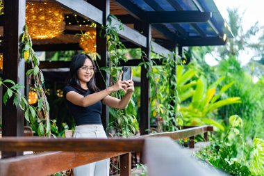 Tropik bir restoran ortamında kendini alan bir kadın. Açık havada ahşap mobilyalar, bereketli yeşillik ve dekoratif ışıklandırma yer almaktadır. Atmosfer rahat ve davetkar.