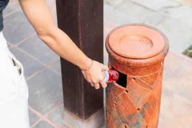 Kırmızı soda kutusunu dekoratif kil çöp kutusuna atan biri. Çöp kutusunun dairesel bir girişi var ve taş kaldırımlarla çevrili olarak dışarıya yerleştirilmiş.