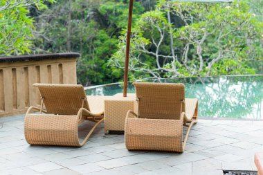 Verandasında şemsiyesi olan iki hasır şezlong yemyeşil manzaraya bakıyor. Sandalyeler cam korkulukların yanında, ağaçların ve doğanın manzarasını sağlıyor..