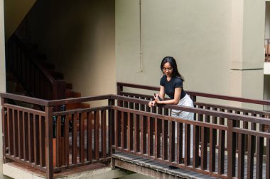 Siyah bluzlu ve beyaz etekli genç bir kadın balkondaki ahşap korkuluklara yaslanıyor. Düşünceli görünüyor, yere bakıyor gibi. Arka planda bir merdiven ve sessiz yeşil bir duvar var.