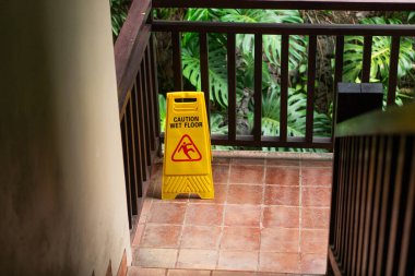 Yarı karanlık bir bölgede, ıslak bir zemini işaret eden parlak sarı bir uyarı işareti. Tabela, kamu alanlarında güvenlik tedbirlerinin vurgulanarak kayan bir kişinin uyarı simgesini içeriyor..