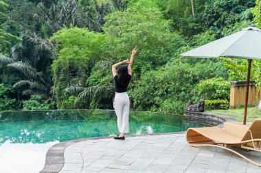 Bir kadın yemyeşil yemyeşil bir havuzun kenarında duruyor. Siyah bir bluz ve beyaz pantolon giyiyor, sakin suya bakıyor.