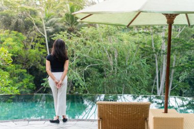 Bir kadın yemyeşil yemyeşil bir havuzun kenarında duruyor. Siyah bir bluz ve beyaz pantolon giyiyor, sakin suya bakıyor.