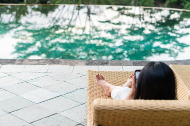Havuzun kenarındaki şezlongda dinlenen akıllı telefonunu kullanan bir kadın. O, siyah bir üst ve beyaz bir pantolon giyiyor, ayakları sandalyede dinleniyor. Sakin havuz suyu.