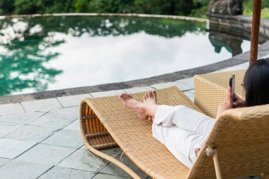 Havuz kenarındaki örülmüş bir paspasın üzerinde dinlenen bir insanın ayaklarının yakından görüntüsü. Ortam sakin ve rahatlatıcı, arka planda bir miktar su yansıması var. Kişi açık renk giyiyor.