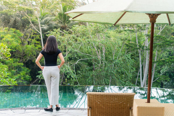 A woman stands by a serene poolside surrounded by lush greenery. She is wearing a black top and white pants, gazing at the tranquil water