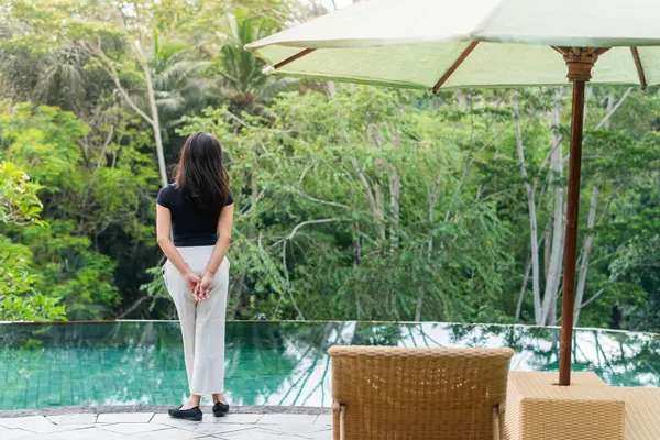 Bir kadın yemyeşil yemyeşil bir havuzun kenarında duruyor. Siyah bir bluz ve beyaz pantolon giyiyor, sakin suya bakıyor.