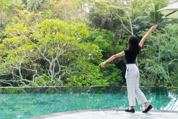 Bir kadın yemyeşil yemyeşil bir havuzun kenarında duruyor. Siyah bir bluz ve beyaz pantolon giyiyor, sakin suya bakıyor.