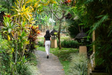 Uzun siyah saçlı genç bir kadın sırtı kameraya dönük duruyor, siyah bir bluz ve beyaz pantolon giyiyor. Canlı tropikal bitkiler ve çiçeklerle dolu yemyeşil bir bahçede.,