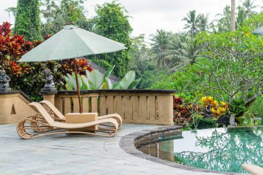 Yeşil şemsiyeli ahşap şezlonglu sakin bir açık havuz. Bereketli tropikal bitkiler ve ağaçlar havuzun etrafını sararak sakin bir atmosfer yaratıyor..