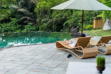 Havuz kenarındaki örülmüş bir paspasın üzerinde dinlenen bir insanın ayaklarının yakından görüntüsü. Ortam sakin ve rahatlatıcı, arka planda bir miktar su yansıması var. Kişi açık renk giyiyor.