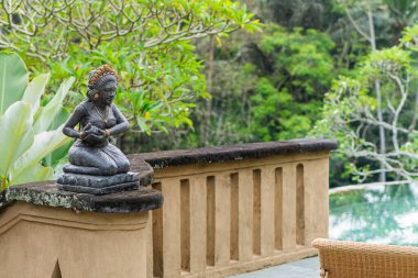 Geleneksel poz veren taştan bir kadın heykeli içeren, bereketli yeşilliklerle çevrili sakin bir açık hava manzarası. Heykel, sakin bir havuz alanına bakan bir taş korkuluğa yerleştirildi.