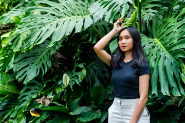 Genç bir kadın büyük yeşil tropikal yaprakların önünde duruyor. Uzun siyah saçları var ve yüksek israf edilmiş beyaz pantolonlu siyah bir bluz giyiyor.