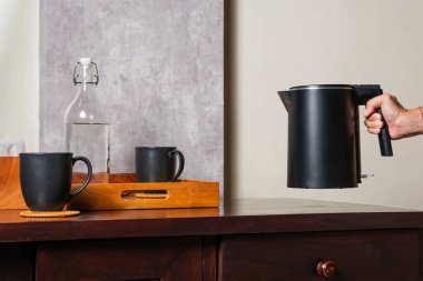 Altında iki siyah bardak olan ahşap bir masanın üzerinde siyah bir elektrikli su ısıtıcısı tutan bir el. Masada, ahşap bir tepsinin yanında suyla dolu bir cam şişe de var..