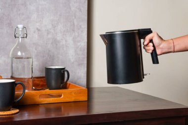 Altında iki siyah bardak olan ahşap bir masanın üzerinde siyah bir elektrikli su ısıtıcısı tutan bir el. Masada, ahşap bir tepsinin yanında suyla dolu bir cam şişe de var..