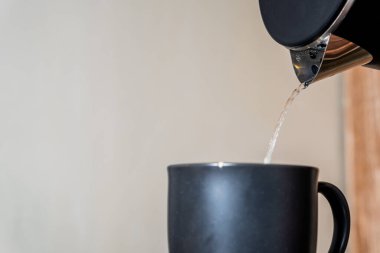 Örgü örülmüş bir bardağa koyulmuş, sıcak su bir çaydanlıktan dökülüyor. Arka planda, bir tahta tepsi, bir bardak ve başka bir siyah bardak var. Ortam çok rahat.