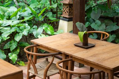 Etrafı yemyeşil bitkilerle çevrili ahşap masalar ve sandalyelerle dolu sakin bir açık hava yemek alanı. Masanın üzerinde ahşaptan yapılmış küçük bir süs eşyası ve bir yaprak duruyor.
