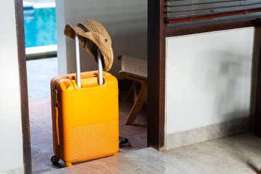 Havuz kenarında turuncu bavullu, şapkalı ve şık giyinmiş bir kadın. Arkaplanda verimli bir yeşillik ve ahşap bir yapı var. Tropik bir tatil ortamı yaratıyor..