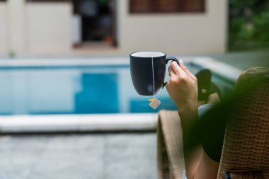 Yüzme havuzunda dinlenen, elinde çay poşeti olan bir fincan çay tutan biri. Sahne sakin, yeşilliğin bulanık arka planı ve berrak bir mavi havuzu var..