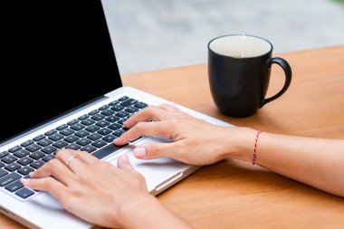 Tahta bir masada dizüstü bilgisayarla otururken elinde sade bir kahve fincanı tutan biri. Laptop ekranı koyu renkli ve arkaplan yeşil renkli bulanık bir açık hava ayarına sahip..