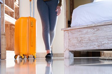 Bir otel odasında tekerlekli parlak sarı bir bavulun yanında duran biri. Bavul yerde dimdik duruyor ve kişi gri tayt giyiyor.