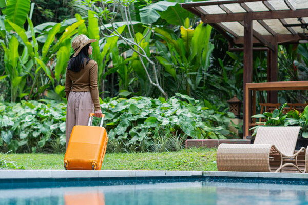 A woman walking by a poolside with an orange suitcase, wearing a hat and stylish outfit. The background features lush greenery and a wooden structure, creating a tropical vacation vibe.