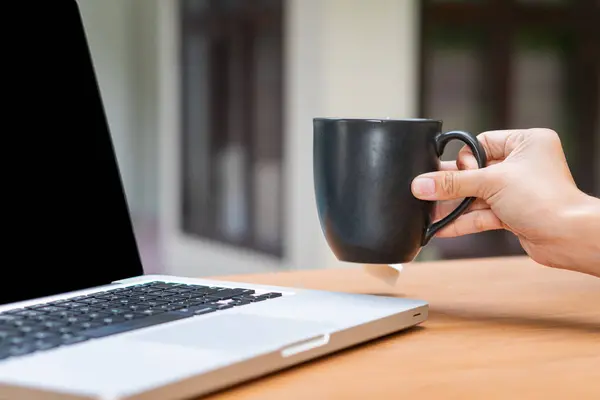 Tahta bir masada dizüstü bilgisayarla otururken elinde sade bir kahve fincanı tutan biri. Laptop ekranı koyu renkli ve arkaplan yeşil renkli bulanık bir açık hava ayarına sahip..