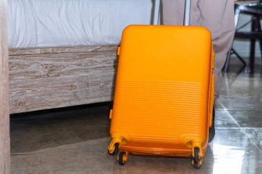 Bir otel odasında tekerlekli parlak sarı bir bavulun yanında duran biri. Bavul yerde dimdik duruyor ve kişi gri tayt giyiyor.