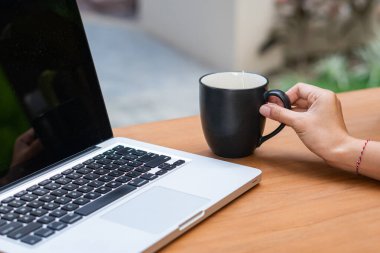 Tahta bir masada dizüstü bilgisayarla otururken elinde sade bir kahve fincanı tutan biri. Laptop ekranı koyu renkli ve arkaplan yeşil renkli bulanık bir açık hava ayarına sahip..