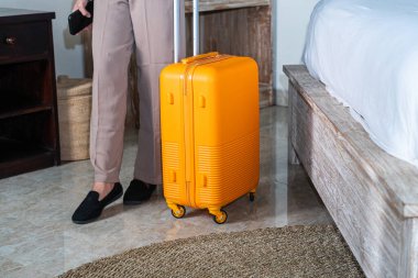 Bir otel odasında tekerlekli parlak sarı bir bavulun yanında duran biri. Bavul yerde dimdik duruyor ve kişi gri tayt giyiyor.